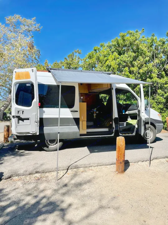 A white & gray converted camper van, excellent for RV rental, with an extended awning & open doors revealing its interior.. Dodge Sprinter Van 2007