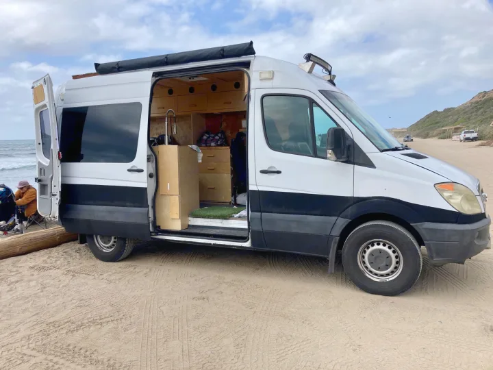 Converted camper van, resembling an RV rental, parked on sandy beach. Open door reveals a custom interior, ready for travel adventures by the ocean.. Dodge Sprinter Van 2007