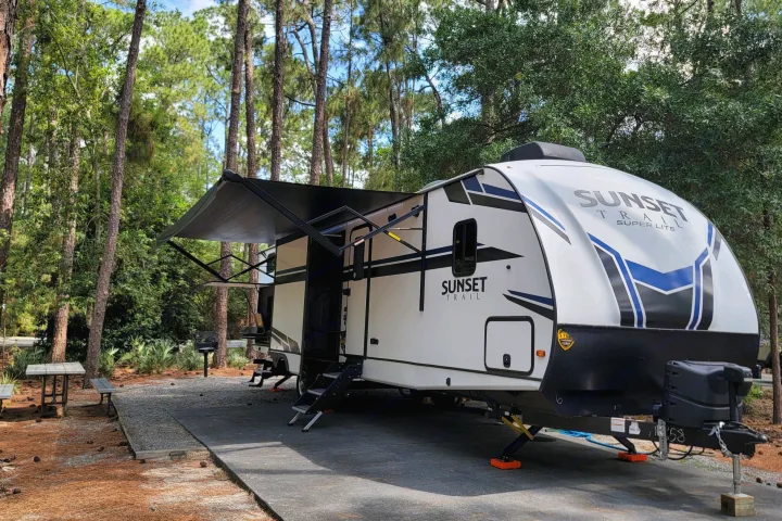Modern rv rental travel trailer parked at a shaded campground site, with its awning extended, a picnic table, and grill.. Crossroads Sunset Trail 2022