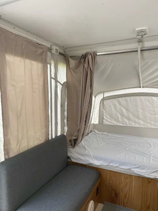 RV rental interior featuring a neatly made bed with white sheets, a gray upholstered sofa, and privacy curtains.. Coleman Destiny Taos 1996