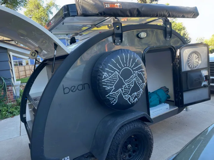 This grey Bean teardrop RV rental shows its rear kitchen galley open, a spare tire with mountain design, and a side door visible, ready for off-road camping.. Bean Mean Bean 2022