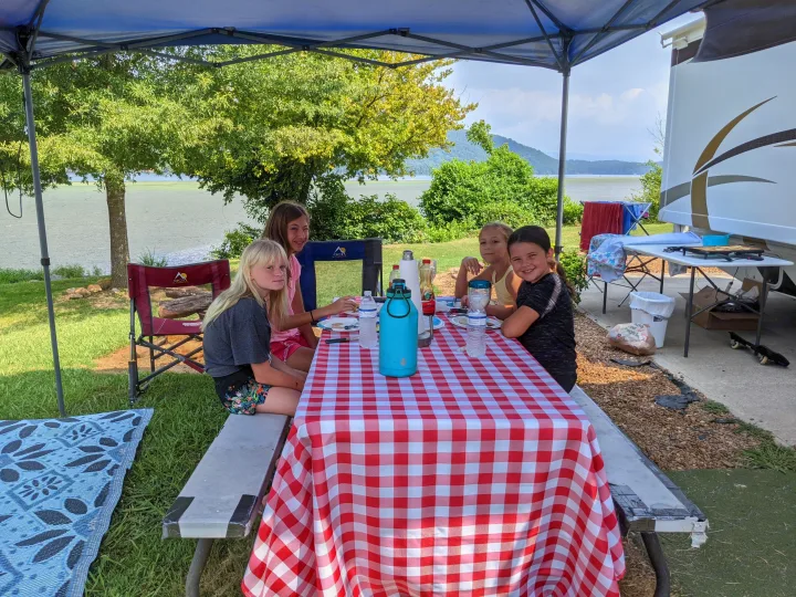 Girls enjoy a picnic meal under a canopy by the lake at an RV rental site, with their RV parked nearby.. Thor Four Winds 2013