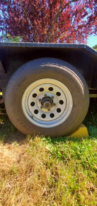 RV rental trailer's durable wheel, tire, and yellow chock securely hold it on the grass, beneath a red-leaf tree.. Eagle flatbed 2019