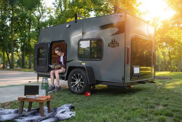 Woman relaxes reading in a compact grey RV rental trailer. Outdoor setup with laptop & blanket in sunny nature.. Superior Lite Camper Deluxe 2022