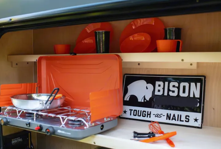 An RV rental's organized kitchenette with a portable stove, pan, orange dishes, and a "BISON" sign. Ready for adventure cooking.. Superior Lite Camper Deluxe 2022