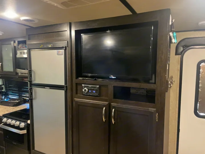 Inside an RV rental: a compact kitchen with fridge, stove, and microwave, plus an entertainment center with TV and stereo.. Grand Design Imagine 2019