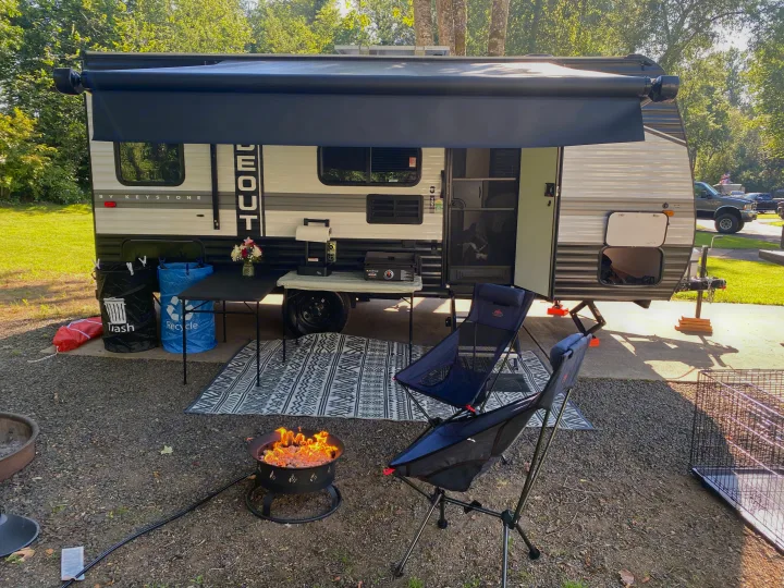 RV rental campsite: travel trailer with extended awning, outdoor seating, tables, and a lit fire pit.. Keystone RV Hideout 2022