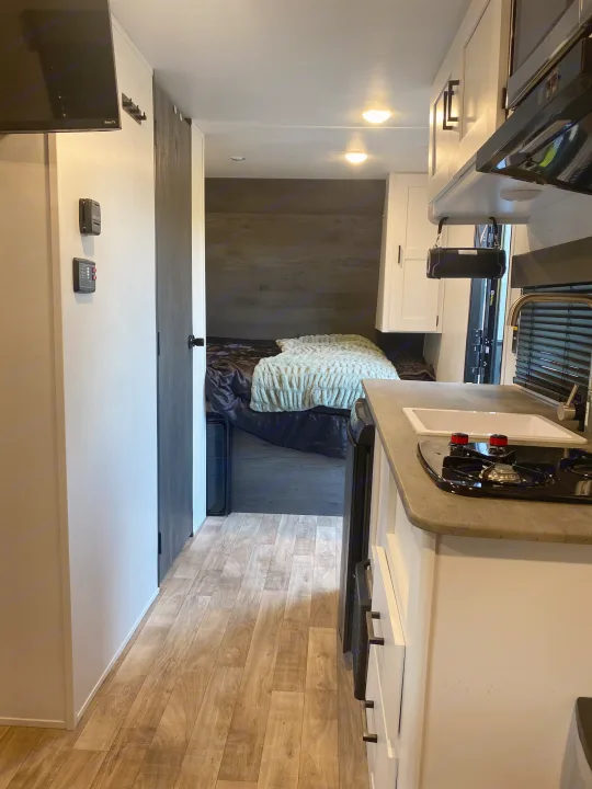 RV rental interior: A functional kitchen with sink & stovetop leads to a cozy bedroom, featuring light wood-look flooring.. Keystone RV Hideout 2022