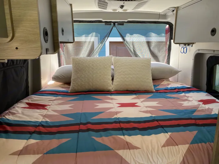 Cozy RV rental sleeping area: Colorful geometric comforter, fluffy pillows, overhead storage, and back window screen.. Winnebago Solis 2021
