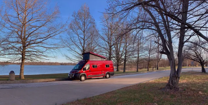 Red pop-top RV rental by a tranquil lake and bare trees. Perfect for nature adventures.. Winnebago Solis 2021