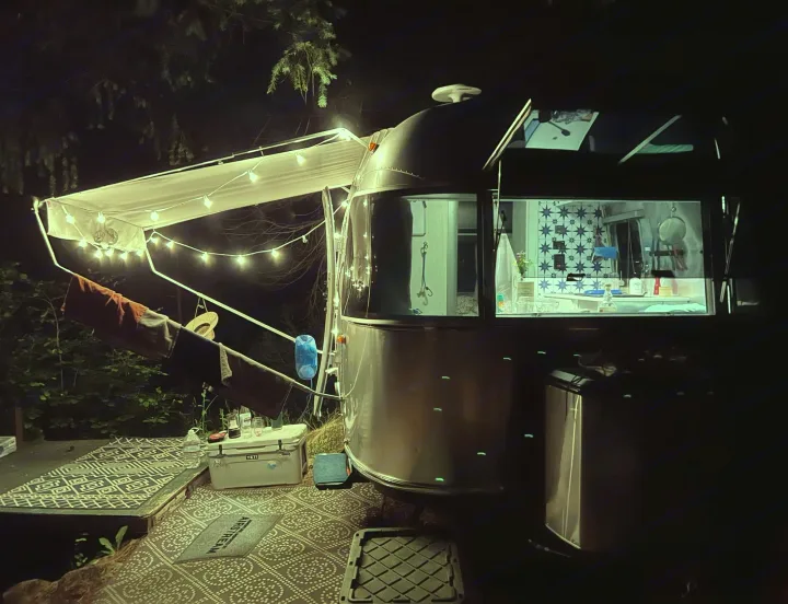Cozy Airstream rv rental at night, with an illuminated awning and patterned outdoor mats, perfect for camping under the stars.. Airstream Bambi 2022