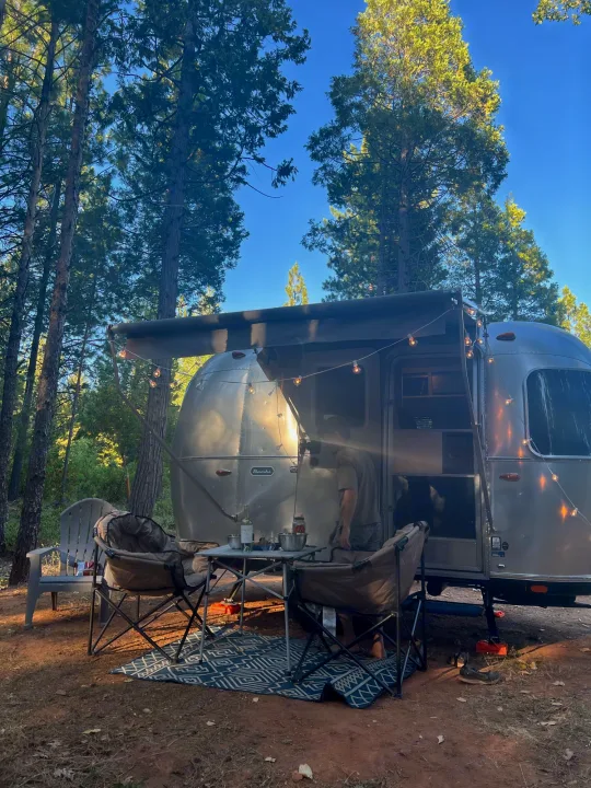 Forest camping with a stylish rv rental. This Airstream is set up with an awning, string lights, and comfy outdoor seating.. Airstream Bambi 2022