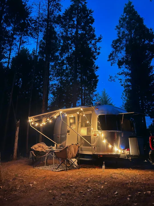 Airstream RV rental glows under string lights at a forest campsite, perfect for night adventures outdoors.. Airstream Bambi 2022
