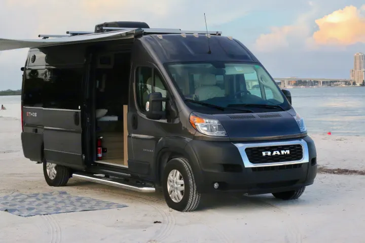 Dark gray Ram camper van, ideal for rv rental, parked on a beach by the water. Awning deployed, side door open.. Entegra Coach Ethos 2021