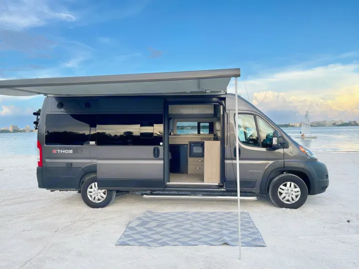 Coastal adventure awaits with this rv rental! A gray camper van with an awning and rug is parked on a sandy beach.. Entegra Coach Ethos 2021
