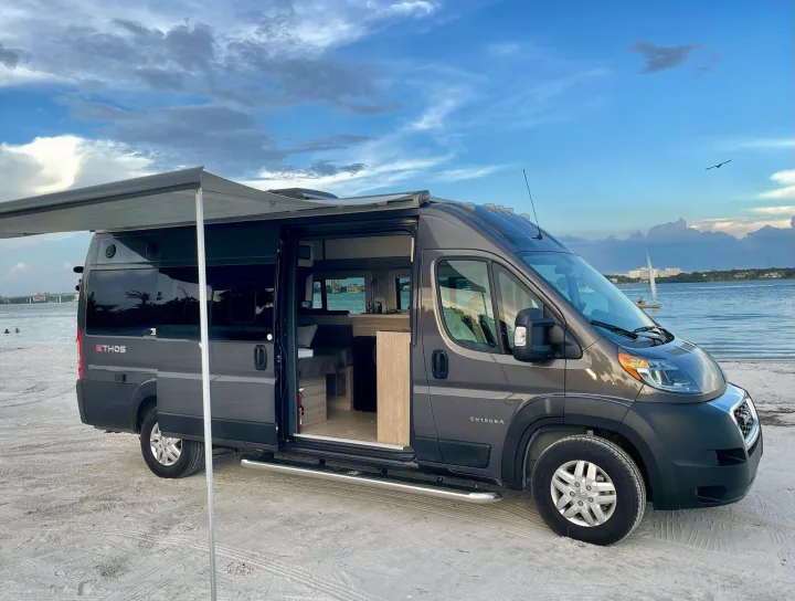 Experience freedom! Dark grey **rv rental** parked on a sandy beach with an open door & extended awning, facing serene waters.. Entegra Coach Ethos 2021