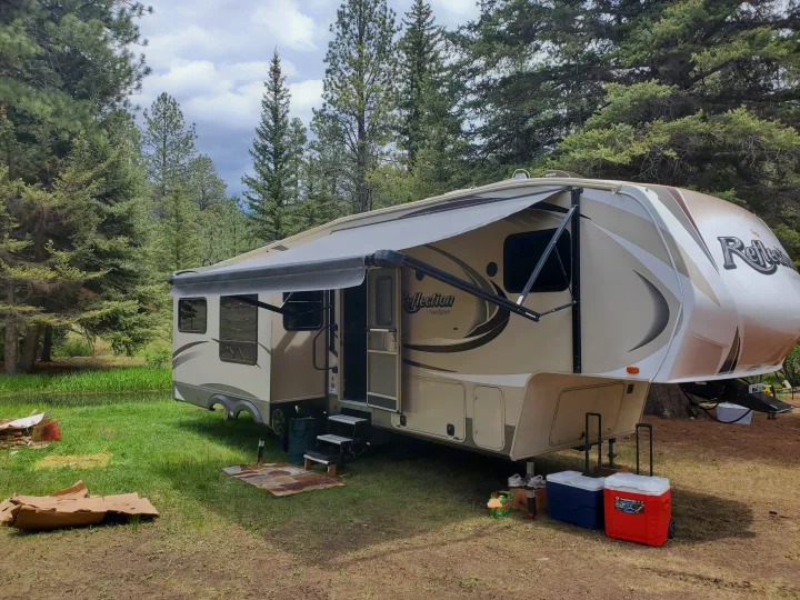 RV rental parked in a tranquil forest campsite, featuring a beige fifth-wheel with awning.. Grand Design Reflection 2015