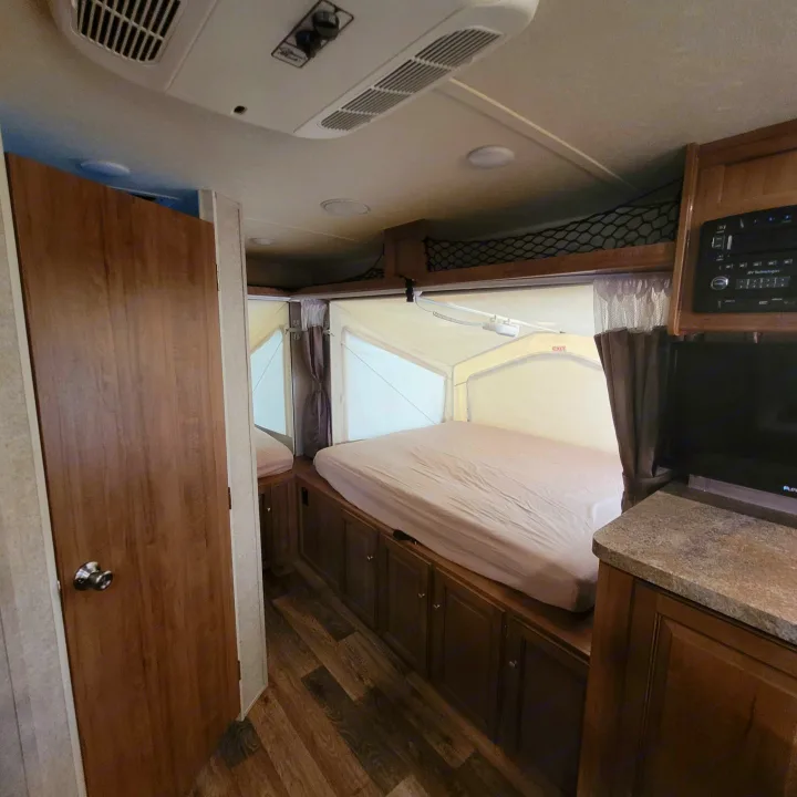 RV rental bedroom area with a pop-out bed, wooden cabinets, door, and ceiling AC unit.. Forest River Rockwood Roo 2017