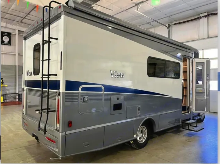 A grey and white 'View' RV rental motorhome is showcased indoors, featuring an open side door and rear access ladder.. Winnebago View 2023