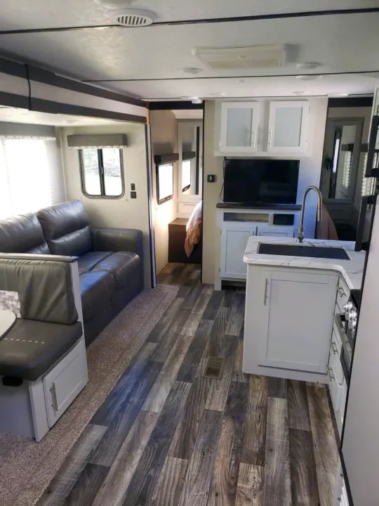 Modern RV rental interior shows a grey sofa, compact kitchen with sink, and wood-look flooring, leading to a back bedroom.. Keystone Bullet 2020