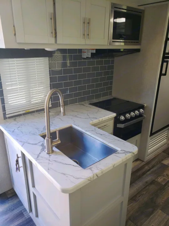 Bright RV rental kitchen with white cabinets, marble-look counters, gray subway tile backsplash, stainless sink, stove, microwave.. Keystone Bullet 2020