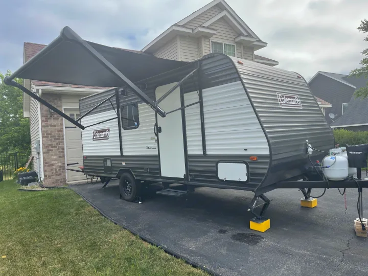 An RV rental, a Coleman travel trailer, is set up on a driveway with its awning extended and stabilizers down.. Dutchmen Coleman Lantern LT 2021