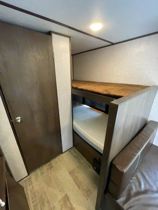 Inside an RV rental: dark wood bunk beds, a closed door, brown leather sofa. Light textured walls, tiled floor.. Dutchmen Coleman Lantern LT 2021