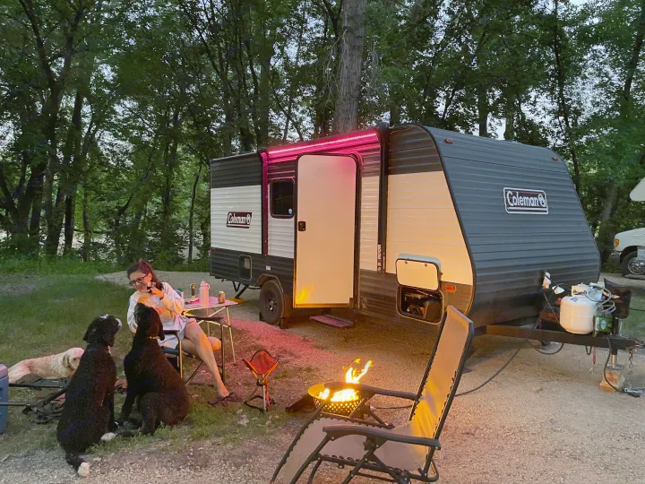 An RV rental glows with red lights at dusk. A person, dogs, and a warm campfire complete this cozy outdoor camping scene.. Dutchmen Coleman Lantern LT 2021