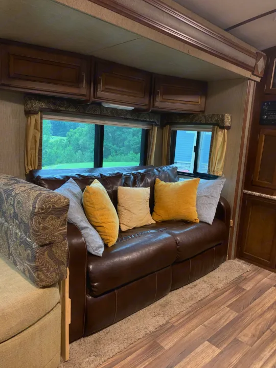 Experience comfort in this RV rental with a plush brown leather sofa, decorative pillows, and expansive windows offering a serene nature view.. Other Outback 2017