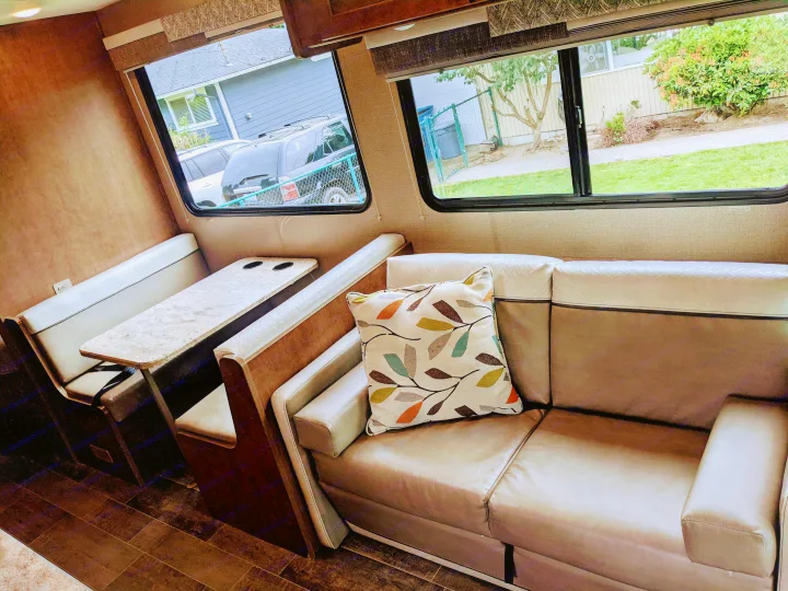 RV rental interior: cozy living space with sofa, patterned pillow, dinette, and large windows showing outdoor views.. Winnebago Outlook 2019