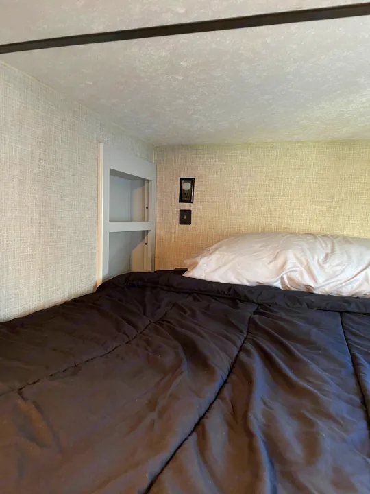 Cozy RV rental bed: bunk with textured walls, built-in shelving, outlets, and a dark comforter.. Keystone Bullet 2020