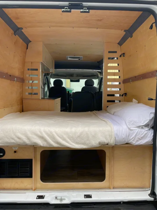Spacious rv rental interior: custom wood camper van with cozy bed & storage, looking forward to front seats.. RAM ProMaster 2022