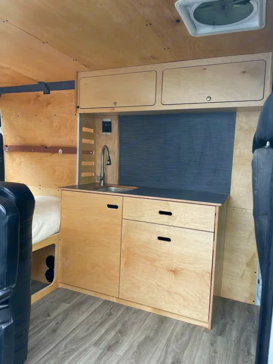 Clean, modern camper van interior, perfect for RV rental. Features a compact kitchen with a sink, wood cabinets & storage.. RAM ProMaster 2022