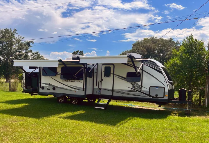 This white & grey travel trailer RV rental, with awnings extended, is parked on green grass under a blue sky. Ready for camping trips.. Jayco White Hawk 2022