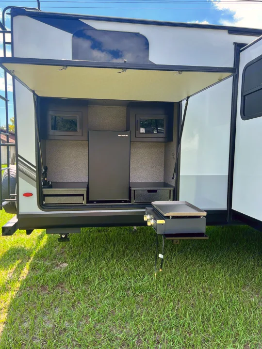 RV rental with rear hatch open, revealing an outdoor kitchen: fridge, sink, and pull-out griddle for cooking, on grassy ground.. Jayco White Hawk 2022