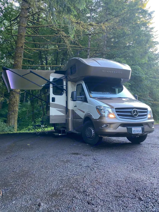 Experience nature with an RV rental! A beige Winnebago motorhome with an extended awning parked at a lush, mossy forest campsite.. Winnebago View 2019