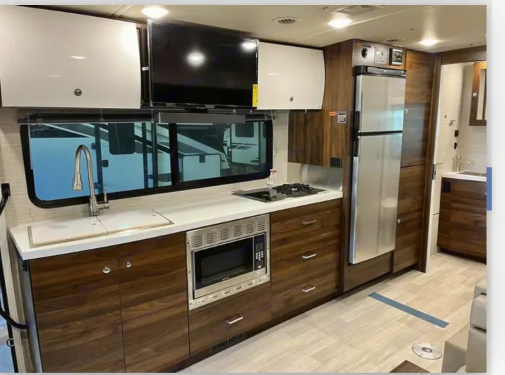 Inside an rv rental, a modern kitchen: sink, cooktop, microwave, and fridge are visible amidst sleek cabinets and a mounted TV.. Winnebago View 2022