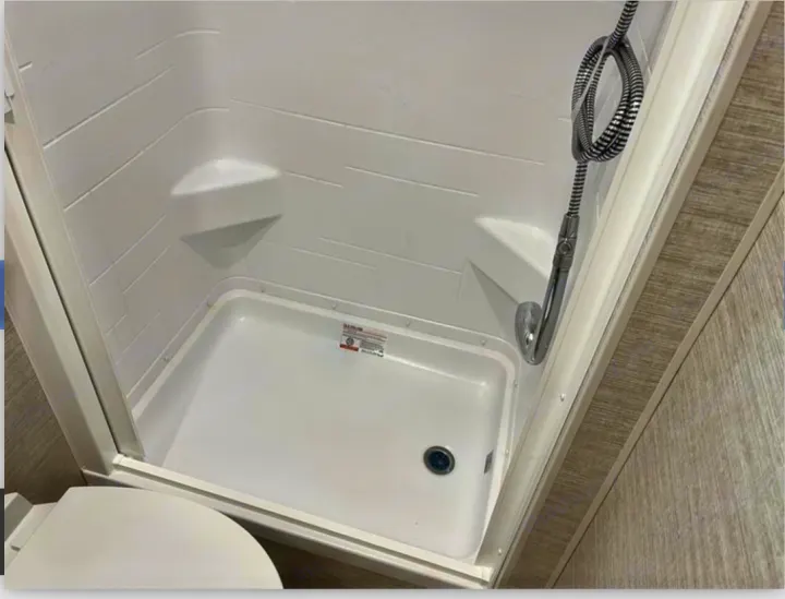 Compact white shower stall in an RV rental, featuring a handheld shower head and corner shelf for convenience.. Winnebago View 2022