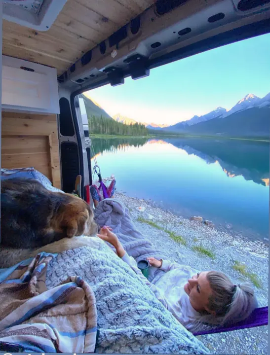 Sunrise over a tranquil lake & mountains from an RV rental. A person sips coffee in a hammock beside their furry friend inside.. Dodge Ram 1500 2020