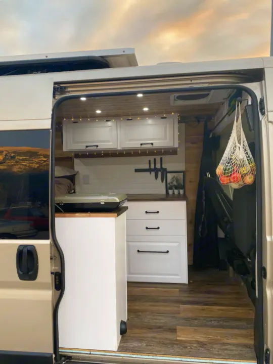 Interior of a modern RV rental camper van, showing a cozy kitchen with white cabinets, wood counters, and warm lighting.. Dodge Ram 1500 2020