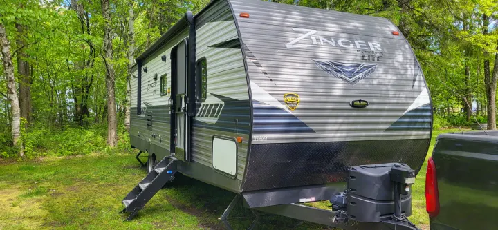 Gray travel trailer RV rental at a shaded campsite, door open with steps down, surrounded by trees.. Crossroads RV Zinger Lite 2020
