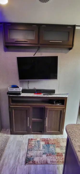 RV rental interior wall: dark cabinets, central TV, and an entertainment setup. A patterned rug hangs on a textured wall.. Crossroads RV Zinger Lite 2020