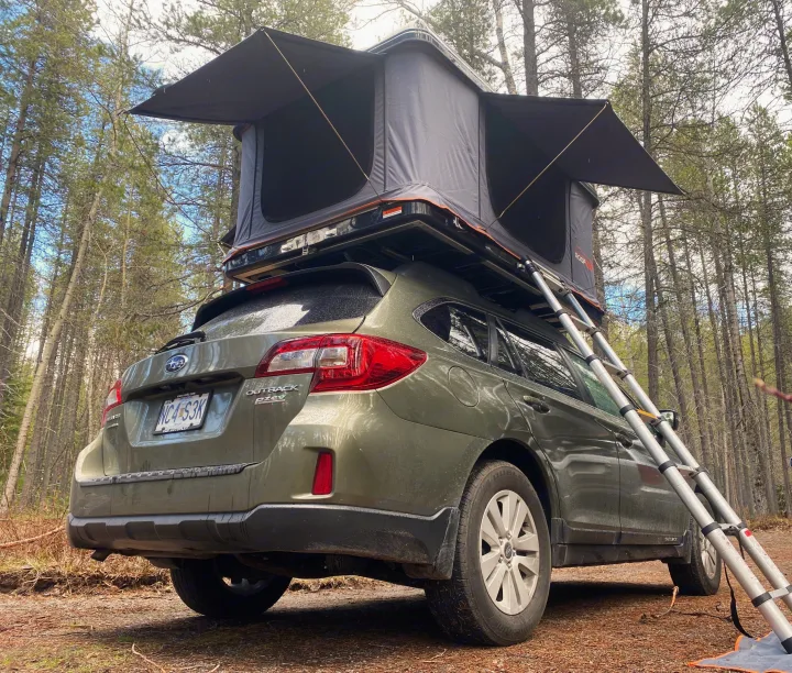 Green Subaru Outback with open rooftop tent & ladder, parked in a forest. Perfect RV rental alternative for camping.. Subaru Outback 2017
