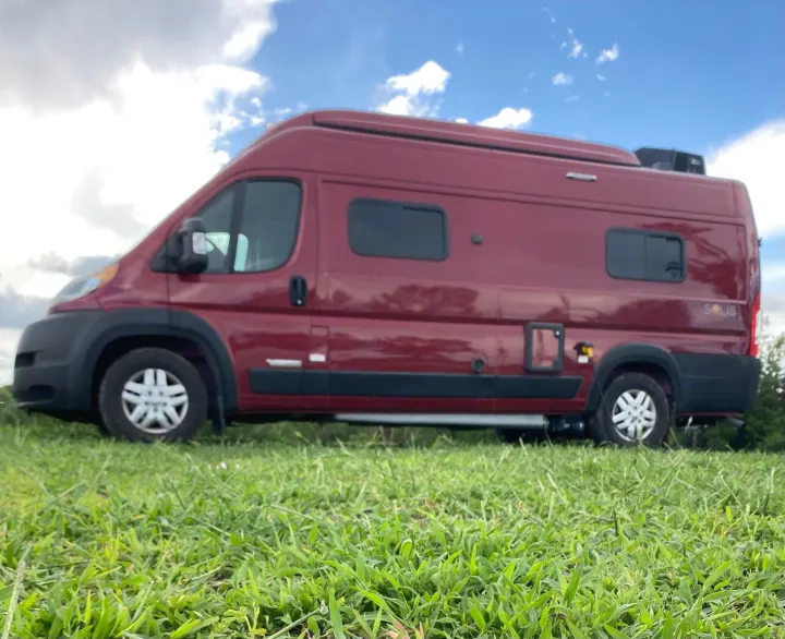 Side view of a red Winnebago Solis camper van, ideal for RV rental, parked on vibrant green grass under a partly cloudy sky.. Winnebago Solis 2021