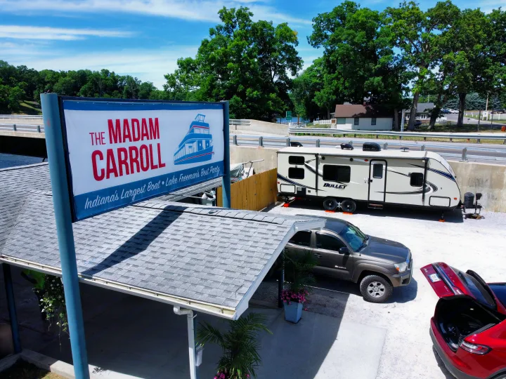 An RV rental travel trailer is parked by "The Madam Carroll" boat sign, alongside other vehicles and green trees.. Keystone Bullet 2017
