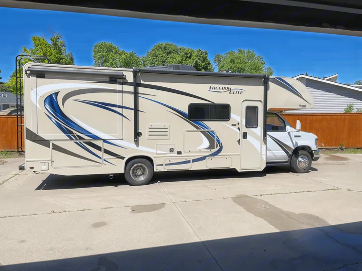 Tan Freedom Elite Class C RV rental, a motorhome with blue/white decals, parked on a sunny driveway for outdoor travel.. Thor Motor Coach Freedom Elite 2019