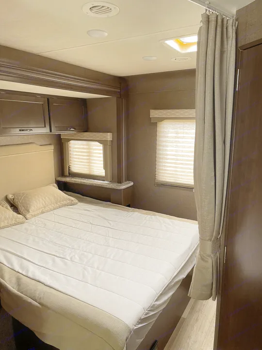 An RV rental bedroom featuring a bed with neutral bedding, two windows with blinds, overhead storage, ceiling lights/vent, and a privacy curtain.. Thor Motor Coach Freedom Elite 2019