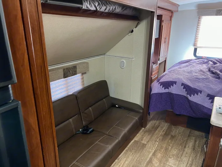 This RV rental interior features a brown sofa, a neatly folded bunk bed, and a comfortable bedroom area with a vibrant purple quilt.. Other Other 2018