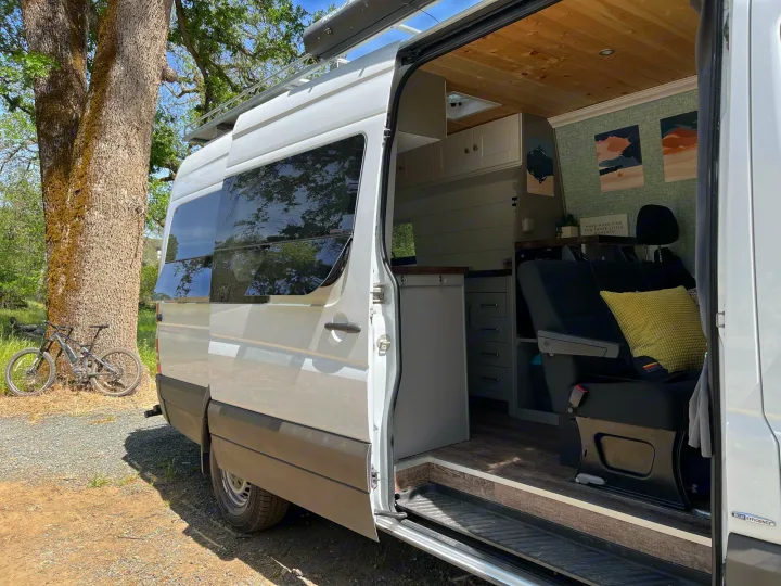 This inviting RV rental shows a white camper van with its door open, revealing a cozy custom interior, parked outdoors with a bike nearby.. Mercedes-Benz Sprinter 2013