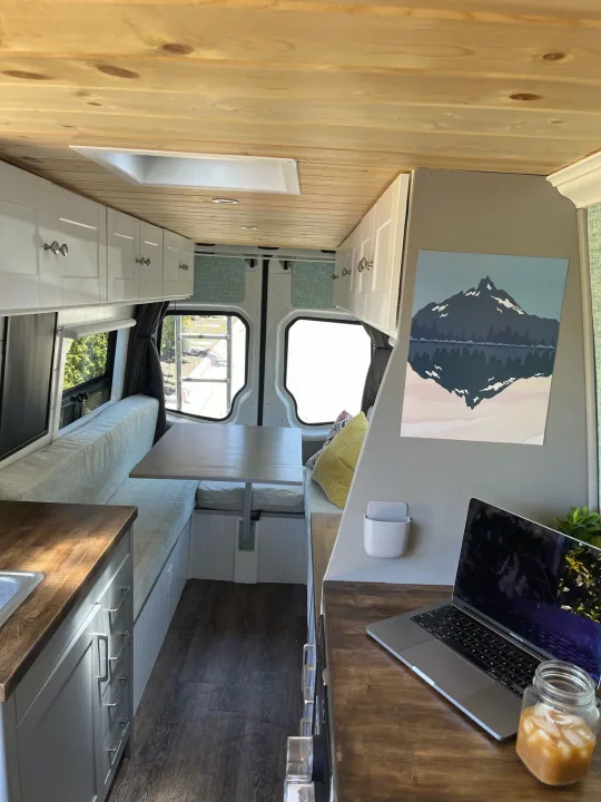 This cozy rv rental interior features a custom wood ceiling, white cabinets, a dining area, and a workspace with a laptop.. Mercedes-Benz Sprinter 2013